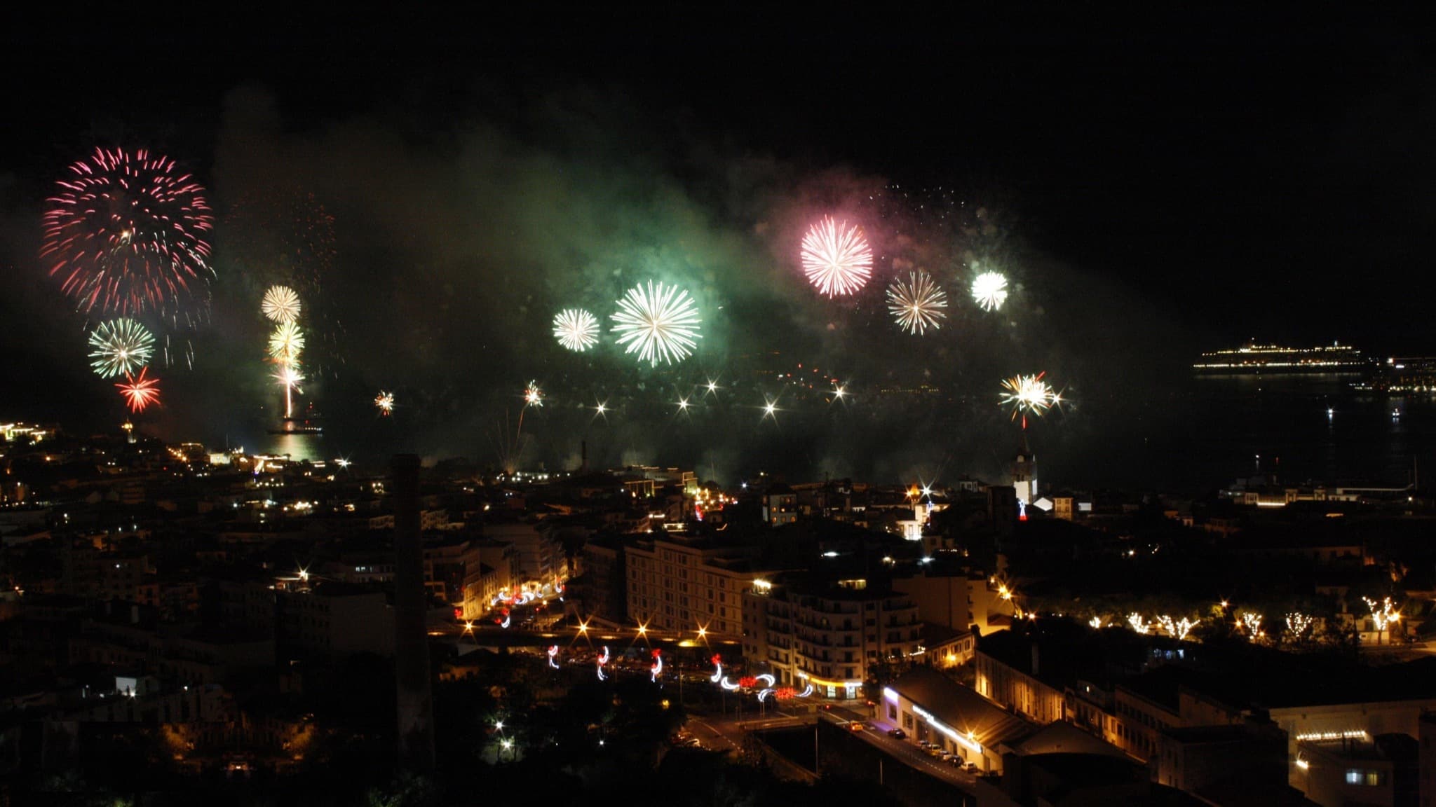 New Year's Eve fireworks firing across the whole bay of Funchal, Madeira, with the lit city below and a cruise ship at anchor