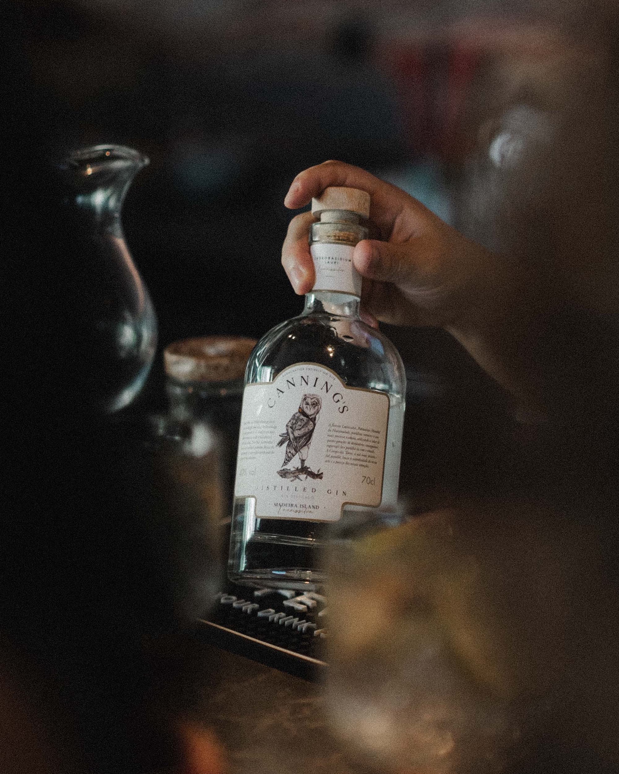 Uma mão a levantar uma garrafa de Canning's pela rolha, num bar em luz quente