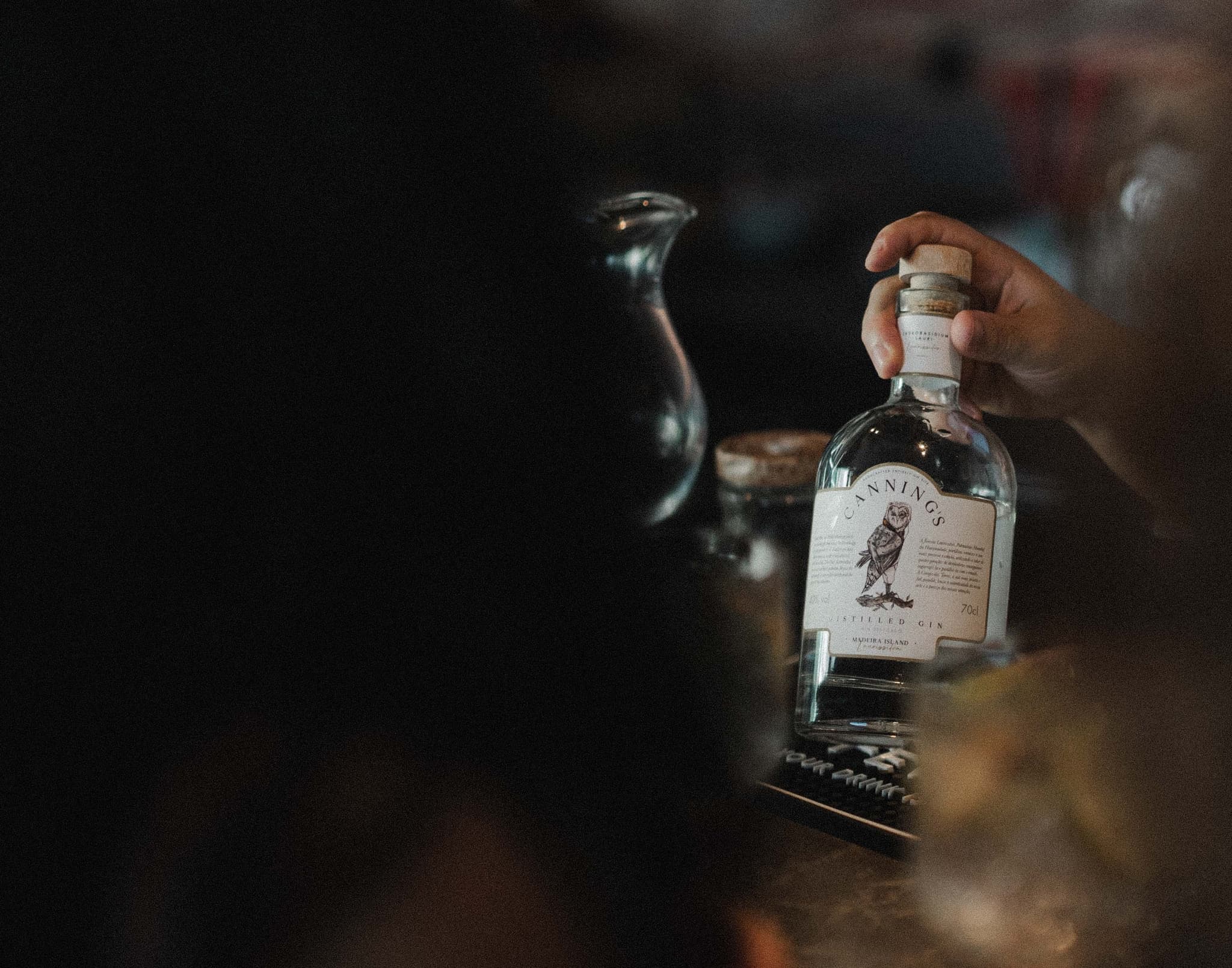 Uma mão a levantar uma garrafa de Canning's pela rolha, num bar em luz quente